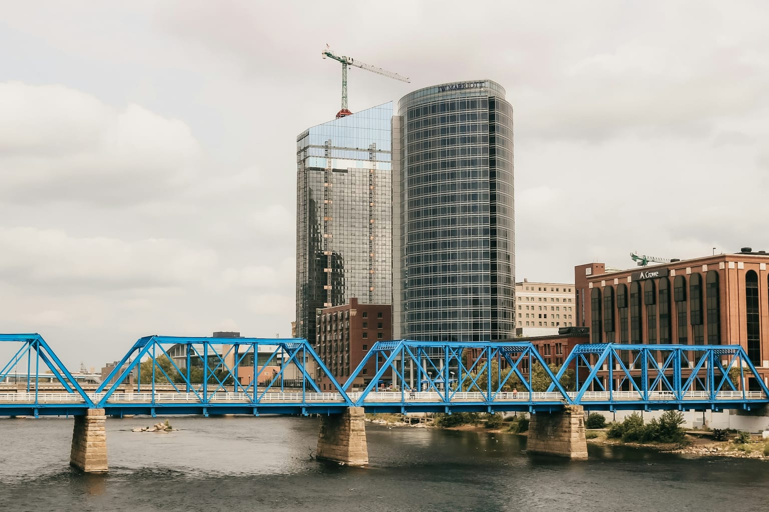 The Blue Bridge, a blue steel truss footbridge, crossing the Grand River in downtown Grand Rapids, with glass towers and brick buildings behind it under an overcast sky.