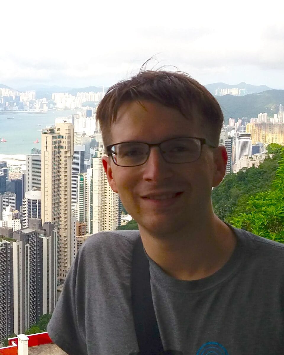 Robby Emmert, smiling, in glasses and a grey T-shirt, with the Hong Kong skyline and harbour behind him.